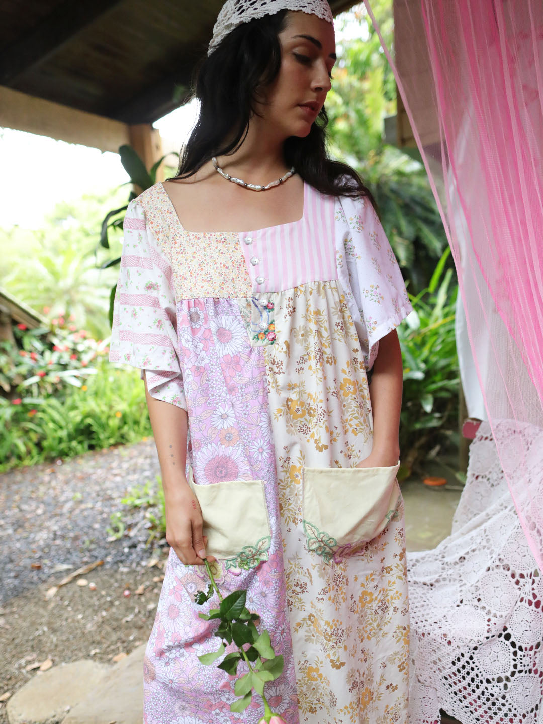 A woman wearing a bohemian patchwork kaftan dress with pink and white floral patterns, standing in a garden setting.