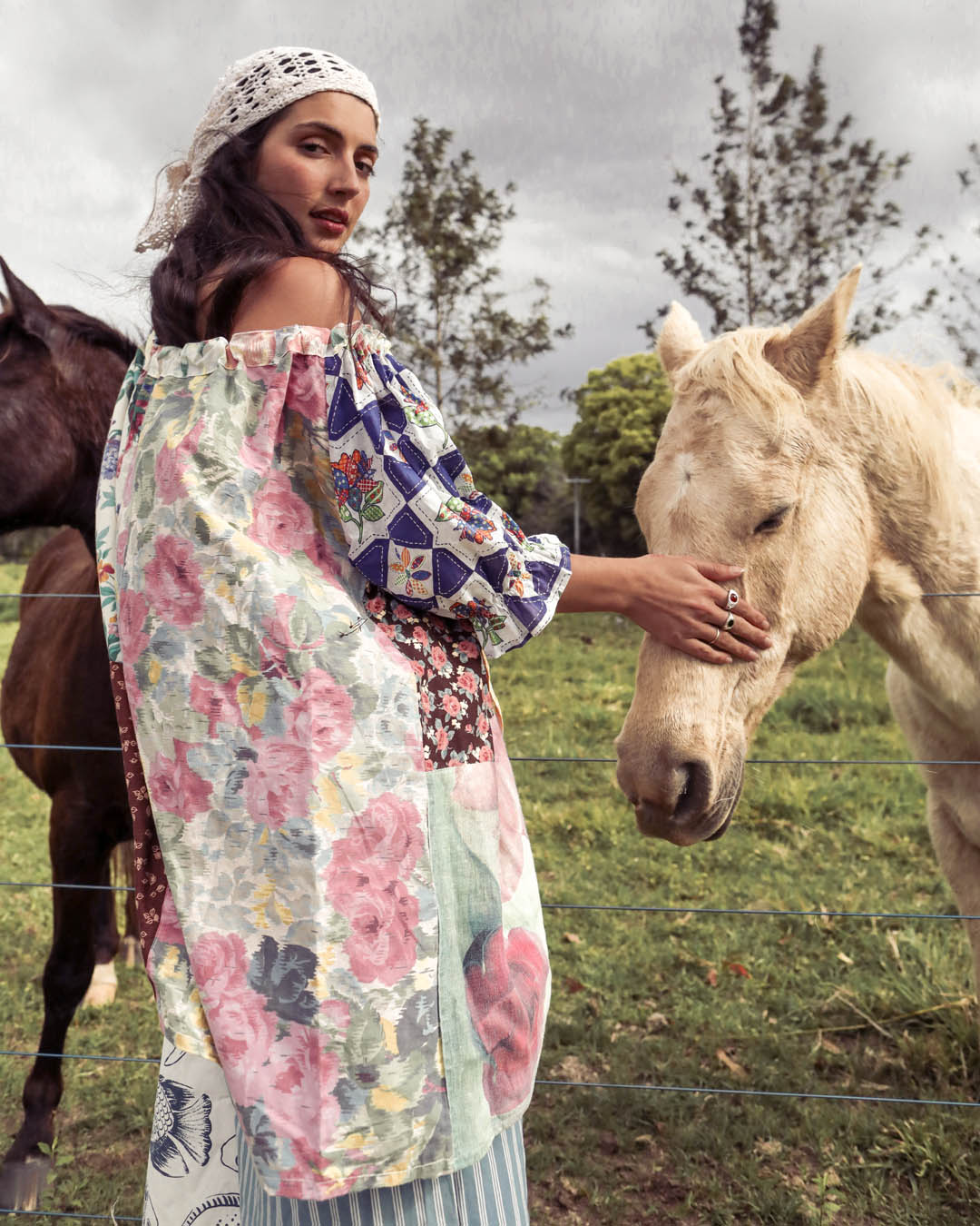 A woman wearing a colorful floral patchwork poncho and a white lace headscarf stands in a grassy field next to a white horse, touching its face. Another dark horse is visible in the background under an overcast sky.