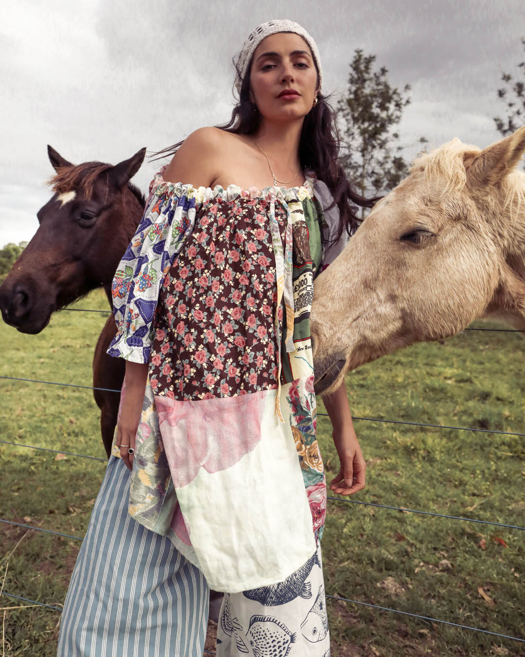 A woman wearing a bohemian patchwork off-the-shoulder top and striped pants stands in a grassy field with two horses, one dark brown and one light tan, under an overcast sky.