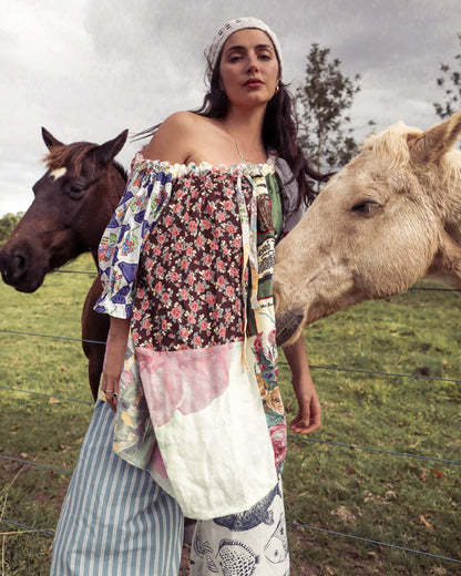 A woman wearing a bohemian patchwork off-the-shoulder top and striped pants stands in a grassy field with two horses, one dark brown and one light tan, under an overcast sky.