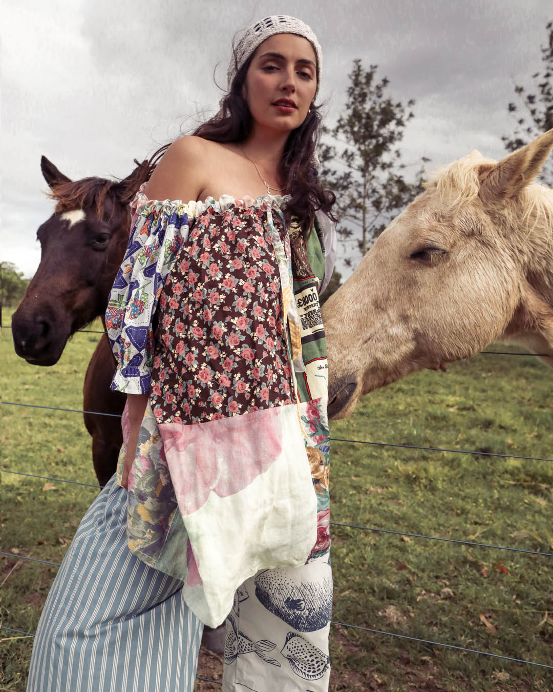 A woman wearing a bohemian-style off-the-shoulder patchwork dress and striped pants stands in a grassy field with two horses, one dark brown and one light beige, under an overcast sky.