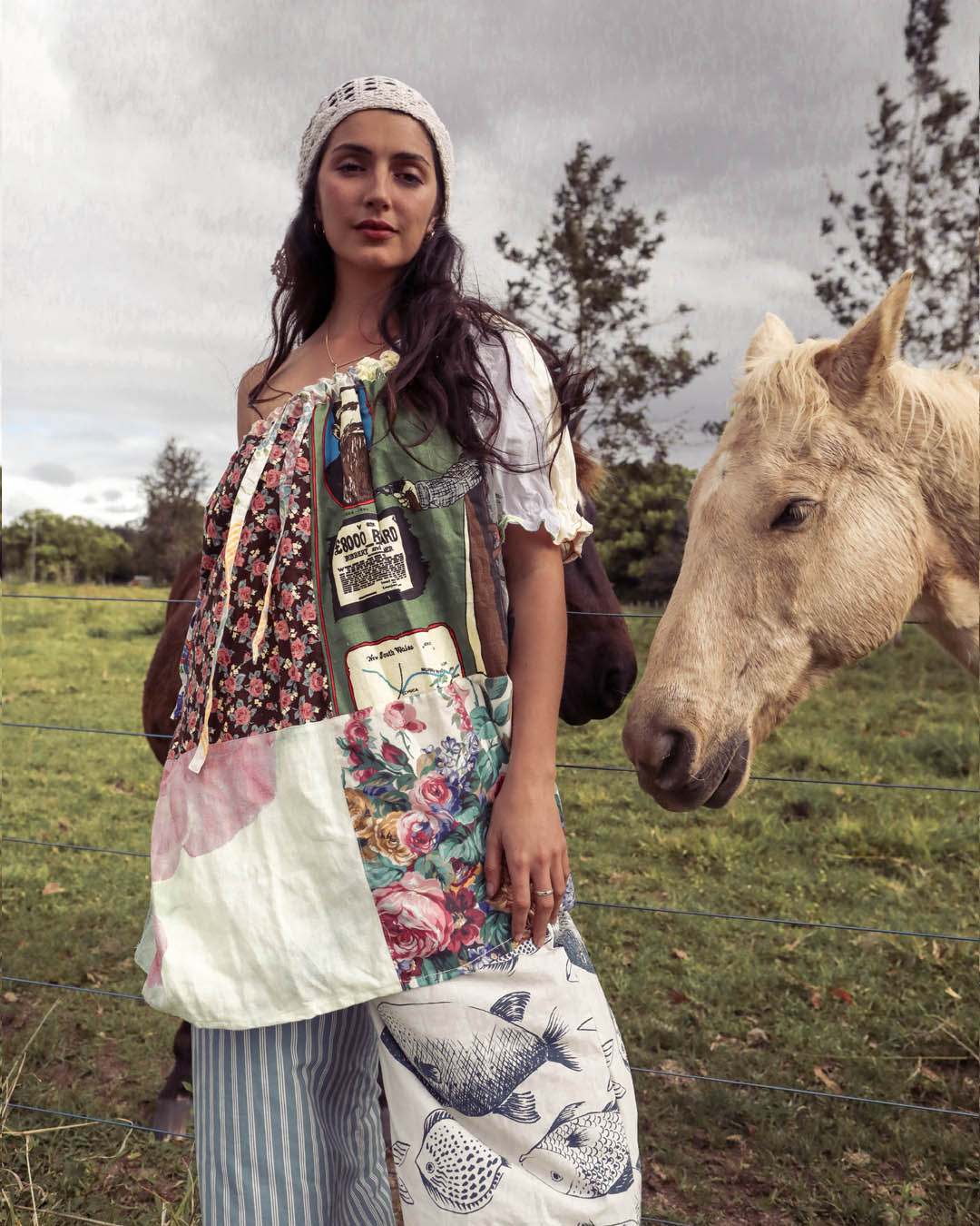 A woman wearing a handmade patchwork quilt dress stands in a grassy field next to a light-colored horse under an overcast sky. The dress features a mix of floral, striped, and quilted panels.