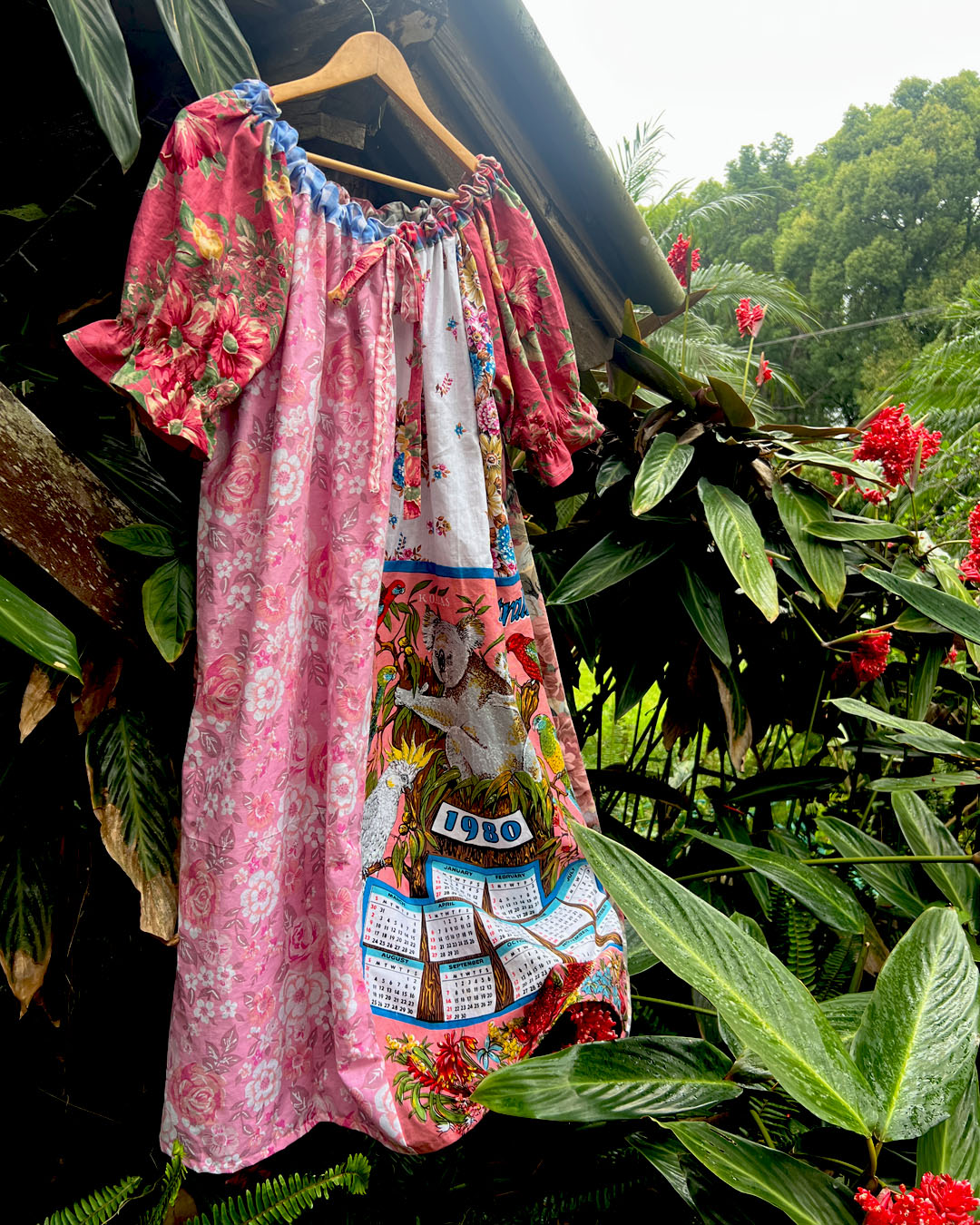 A multicolor bohemian-style smock dress with pink floral patterns and a central illustration of a koala, Australian native birds and calendar for the year 1980, hanging on a wooden hanger against a lush green tropical foliage background.