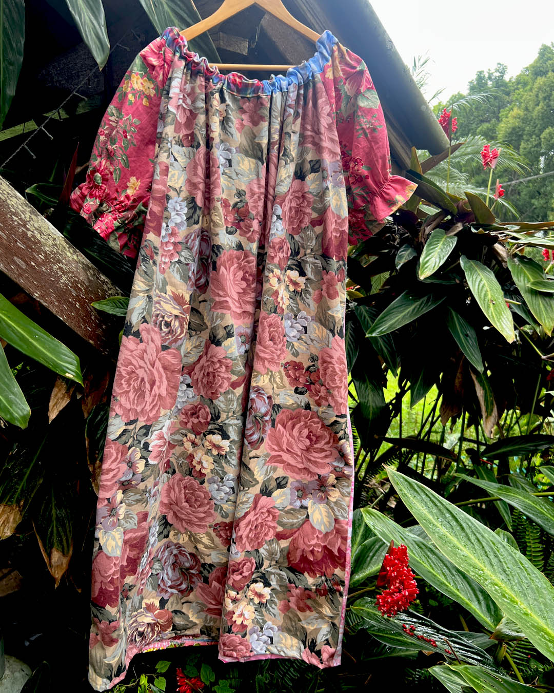 A short-sleeve floral dress with a gathered neckline and ruffled sleeves, hanging on a wooden hanger against a backdrop of tropical green foliage and red flowers.