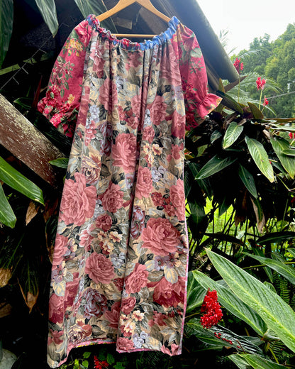 A short-sleeve floral dress with a gathered neckline and ruffled sleeves, hanging on a wooden hanger against a backdrop of tropical green foliage and red flowers.