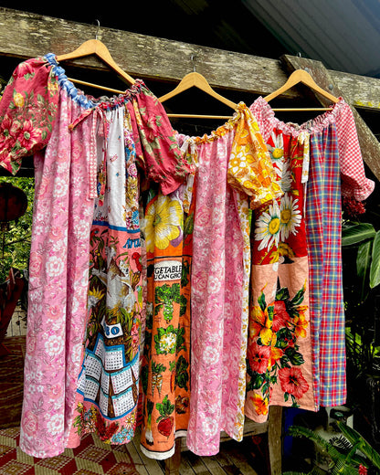 A group of colorful vintage-style patchwork dresses hanging on wooden hangers outdoors against a rustic wooden fence. The dresses feature various patterns including florals, plaid, and vegetable prints in pink, orange, yellow, red, and blue.