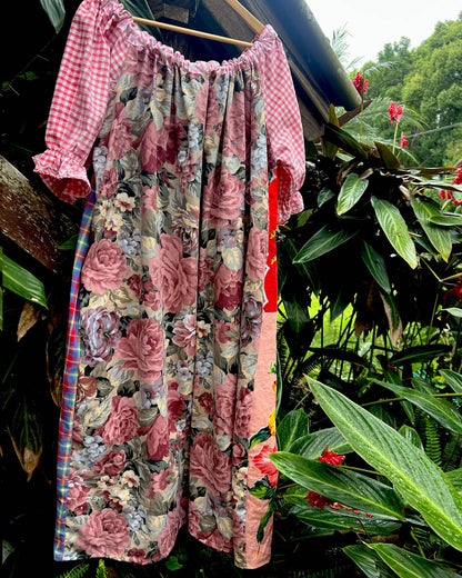 A short-sleeve dress with a pink and white gingham bodice and a floral skirt featuring pink roses, hanging outdoors against a backdrop of lush green tropical plants.