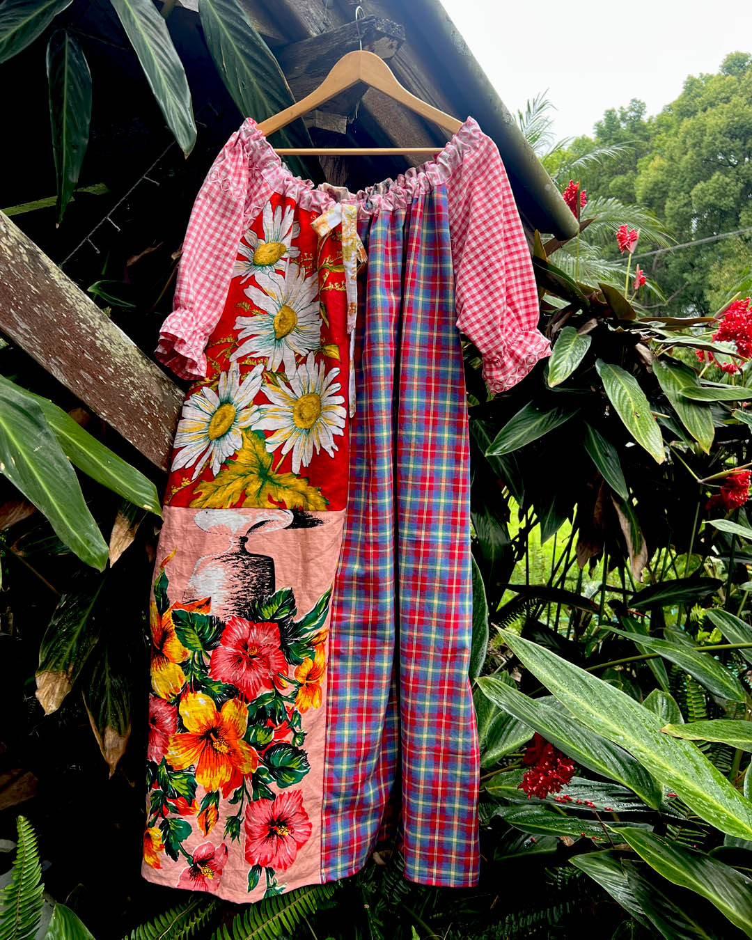 A patchwork-style cotton one size, one of a kind smock dress hanging on a wooden hanger, featuring a red gingham bodice, floral daisy panels, hibiscus flowers, and a red/blue plaid cotton photographed outdoors against tropical green foliage.