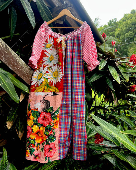 A patchwork-style cotton one size, one of a kind smock dress hanging on a wooden hanger, featuring a red gingham bodice, floral daisy panels, hibiscus flowers, and a red/blue plaid cotton photographed outdoors against tropical green foliage.