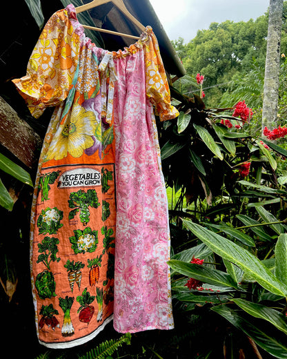 One size, one of a kind patchwork women's smock dress hanging on a wooden hanger outdoors. Drawstring neckline, puff sleeves in yellow, orange, and floral print with heirloom vegetable tea towel linen.