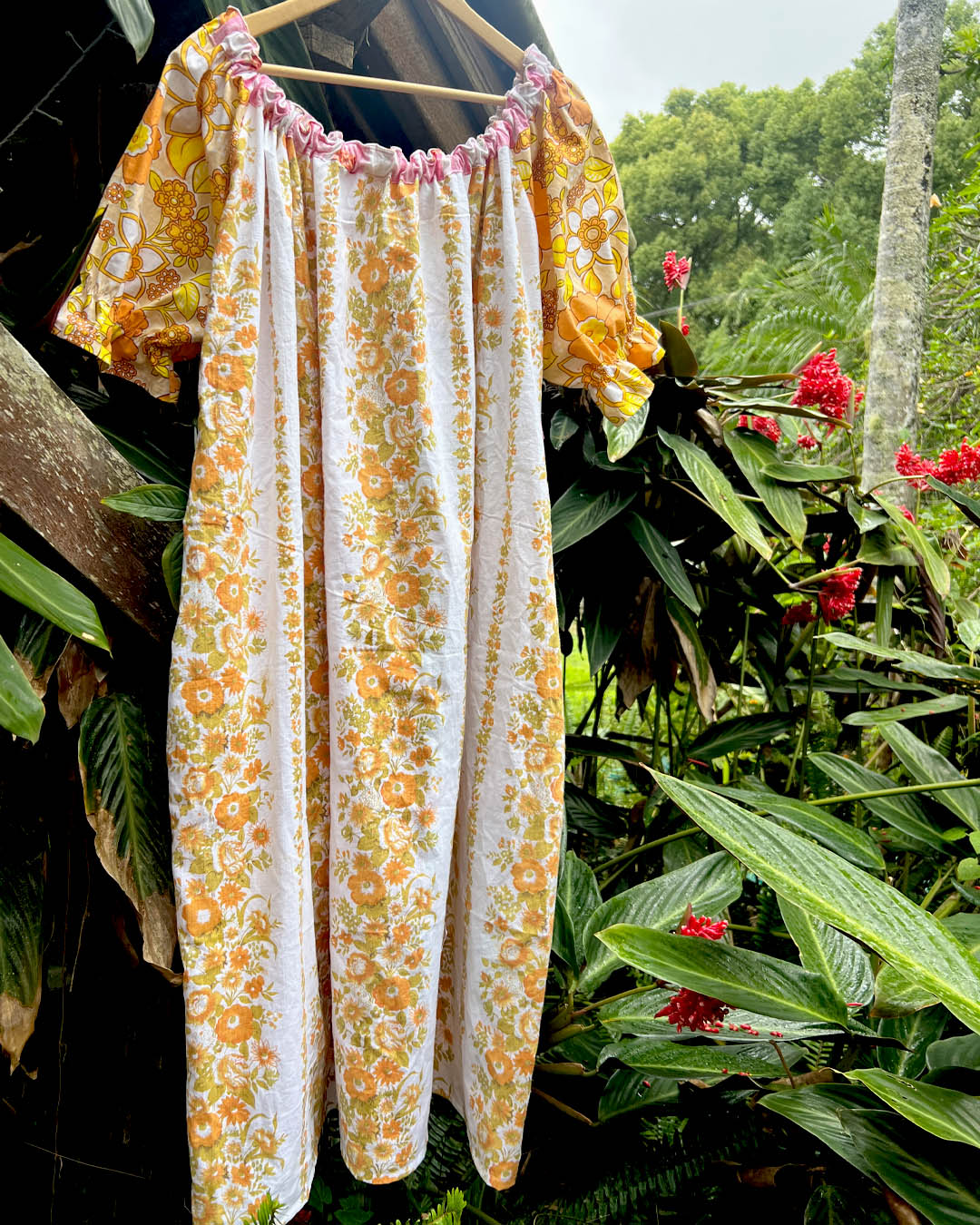 A short-sleeve dress with a yellow, orange, and white floral print hanging on a wooden hanger against a lush green garden background with red flowers.