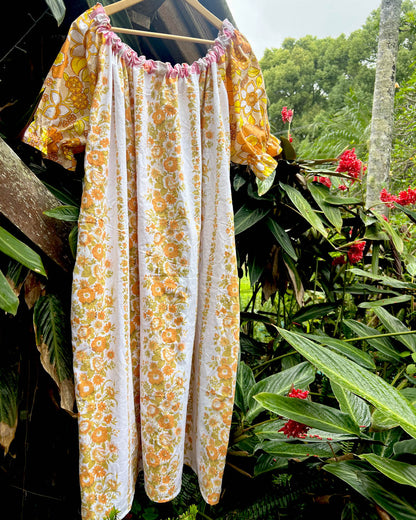 A short-sleeve dress with a yellow, orange, and white floral print hanging on a wooden hanger against a lush green garden background with red flowers.