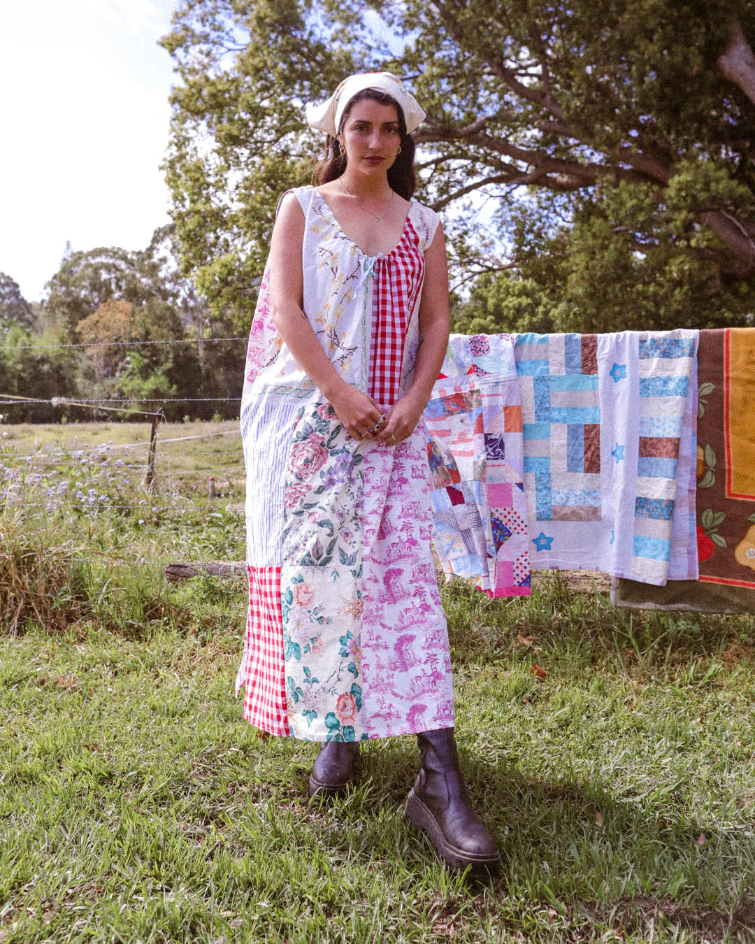 A woman stands outdoors in a grassy yard wearing a long patchwork dress made from multiple colorful fabrics, with a white headband and dark brown boots. A quilted blanket hangs on a clothesline in the background among trees.