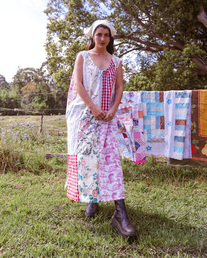 A woman stands outdoors in a grassy yard wearing a long patchwork dress made from multiple colorful fabrics, with a white headband and dark brown boots. A quilted blanket hangs on a clothesline in the background among trees.