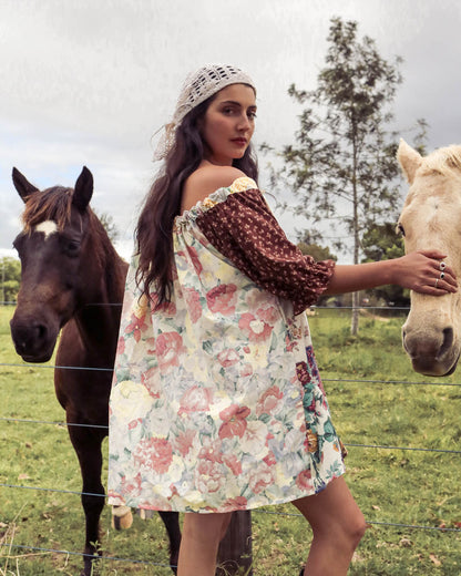 A woman wearing a bohemian-style floral dress and crochet headscarf stands in a grassy field with two horses behind her, looking back over her shoulder.
