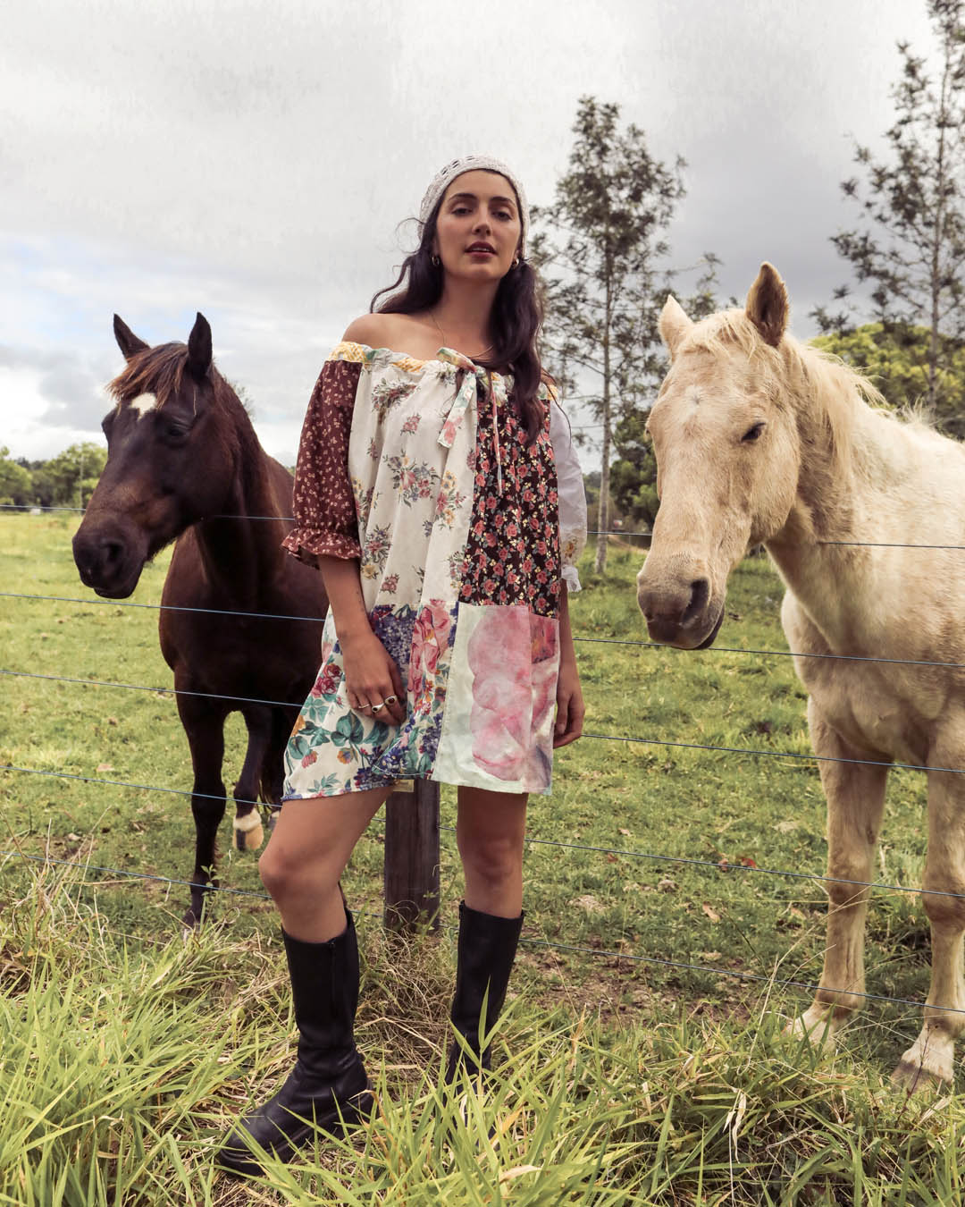 A woman stands in a grassy field between two horses, wearing a patchwork off-the-shoulder dress with floral and patterned panels, black knee-high boots, and a white headband.