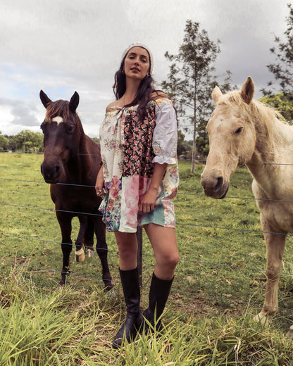 A woman stands in a grassy field between two horses, wearing a bohemian patchwork dress with floral and paisley prints, a brown leather crossbody, and black knee-high boots.