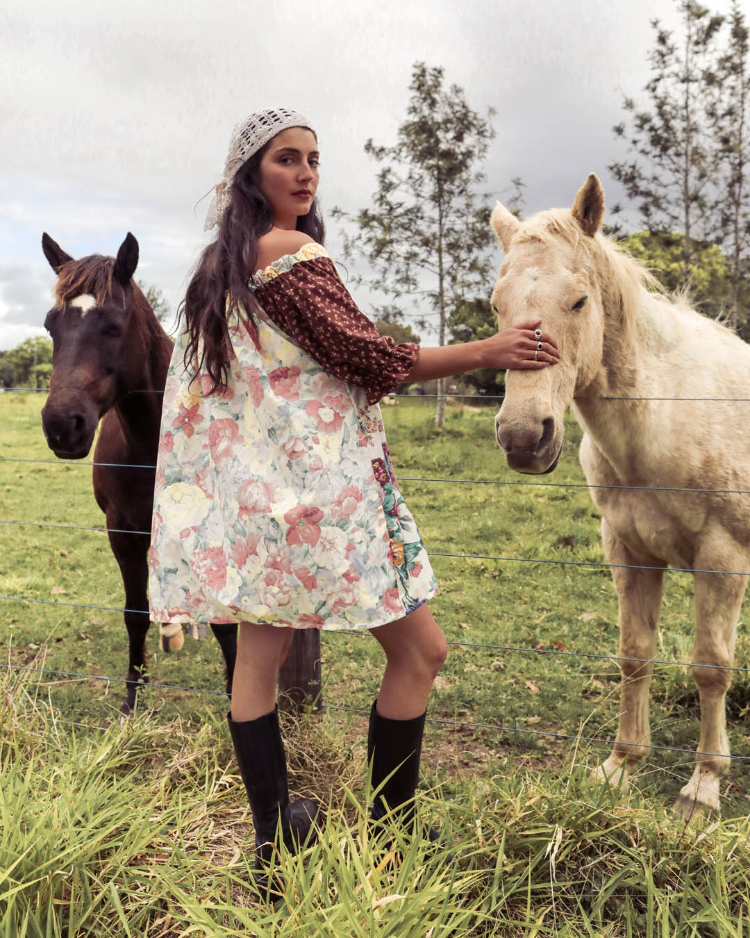 A woman in a floral off-the-shoulder dress and knit hat stands in a grassy field between two horses, touching the cream horse's face. The scene has a vintage, filtered look with muted colors.