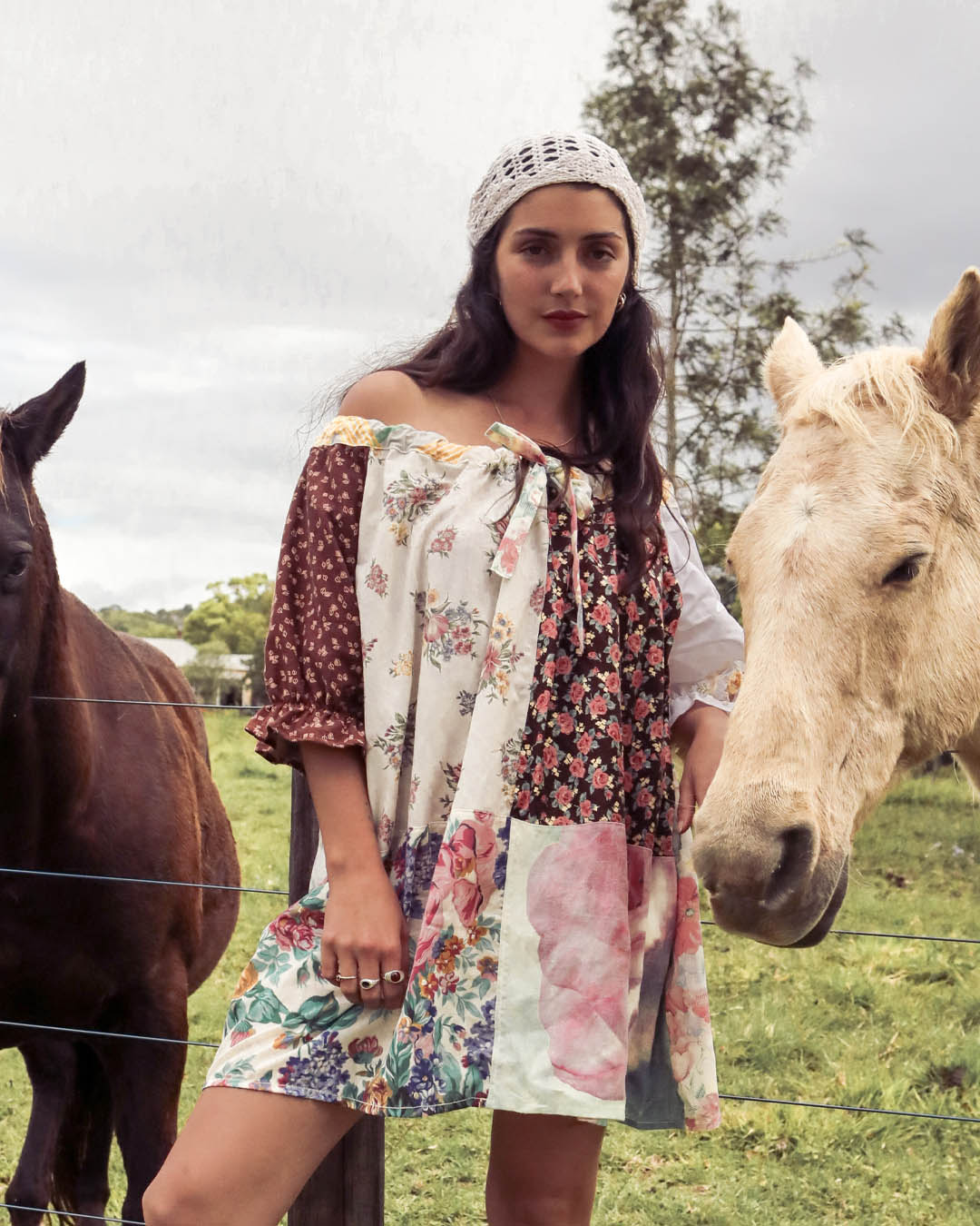 A woman wearing a bohemian off-the-shoulder patchwork mini dress and a white knit beanie stands outdoors in a grassy field with two horses on either side of her.