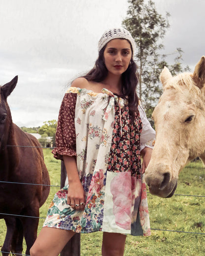 A woman wearing a bohemian off-the-shoulder patchwork mini dress and a white knit beanie stands outdoors in a grassy field with two horses on either side of her.