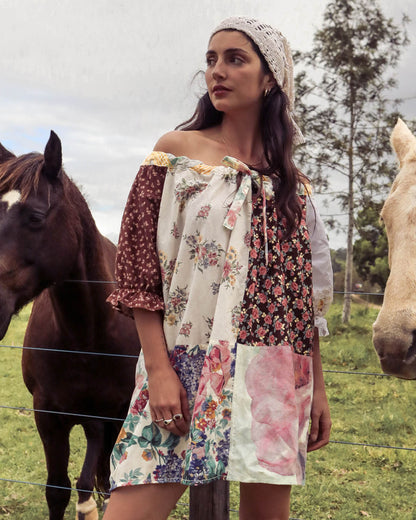 A woman wearing a bohemian off-the-shoulder patchwork dress and a white crochet beanie stands in a grassy field with horses on either side of her.
