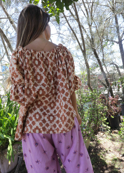 Woman wearing an ochre handblock patterned top and purple pants standing outdoors with trees and plants in the background. A perfect Christmas Fashion Outfit and Black Friday Sale