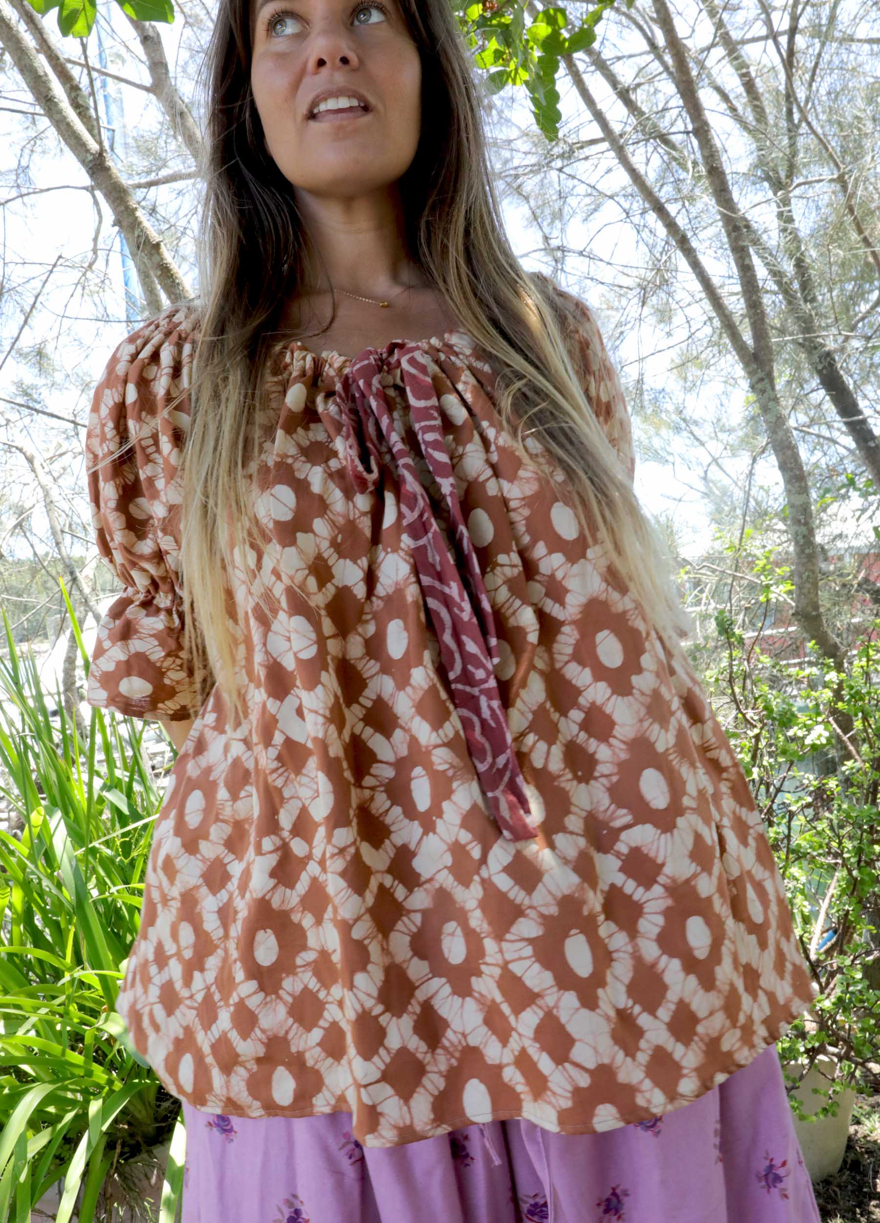 Woman wearing an ochre handblock patterned top and purple pants standing outdoors with trees and plants in the background. A perfect Christmas Fashion Outfit and Black Friday Sale