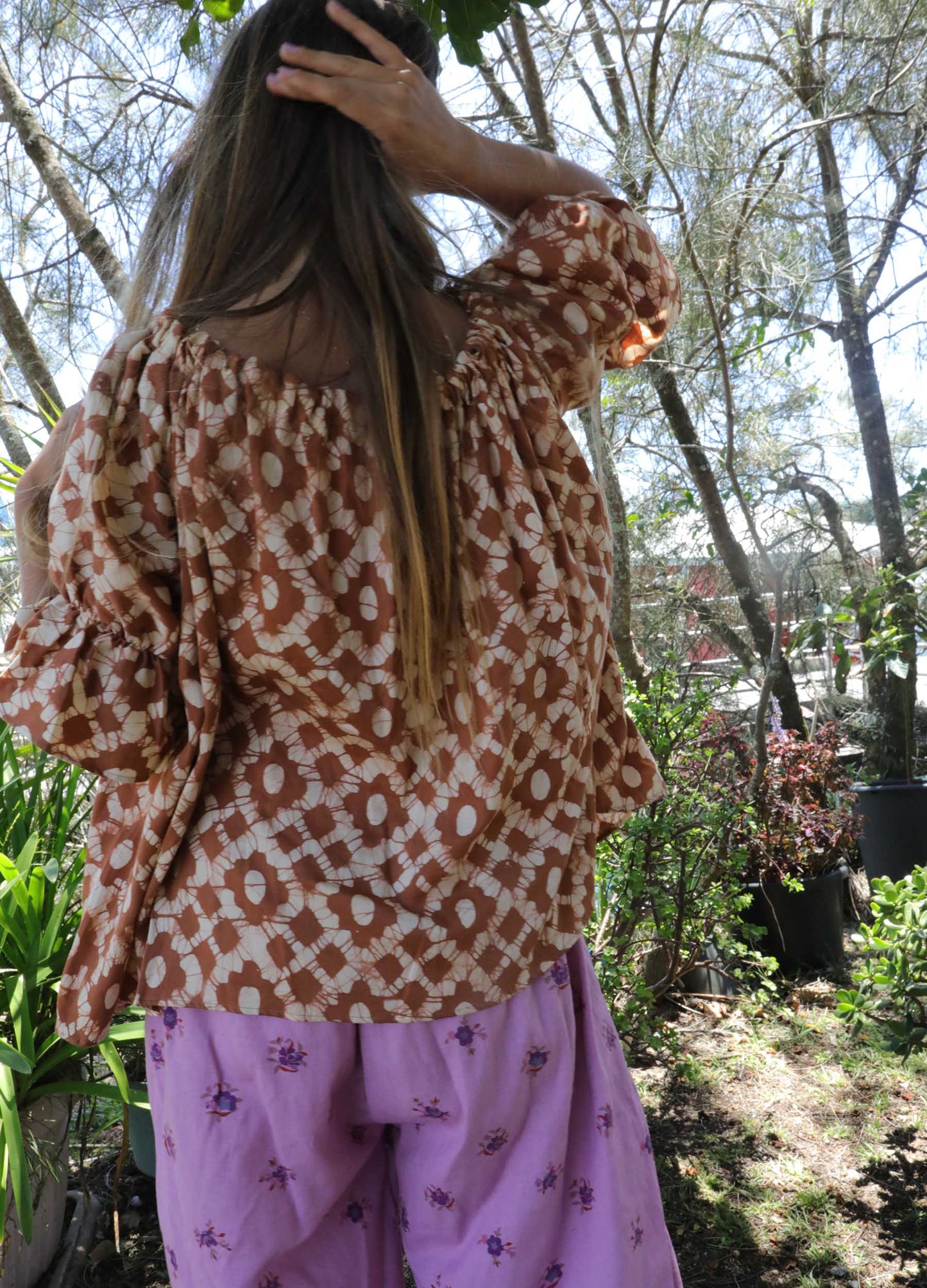 Woman wearing an ochre handblock patterned top and purple pants standing outdoors with trees and plants in the background. A perfect Christmas Fashion Outfit and Black Friday Sale