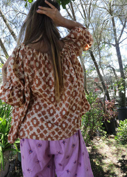 Woman wearing an ochre handblock patterned top and purple pants standing outdoors with trees and plants in the background. A perfect Christmas Fashion Outfit and Black Friday Sale