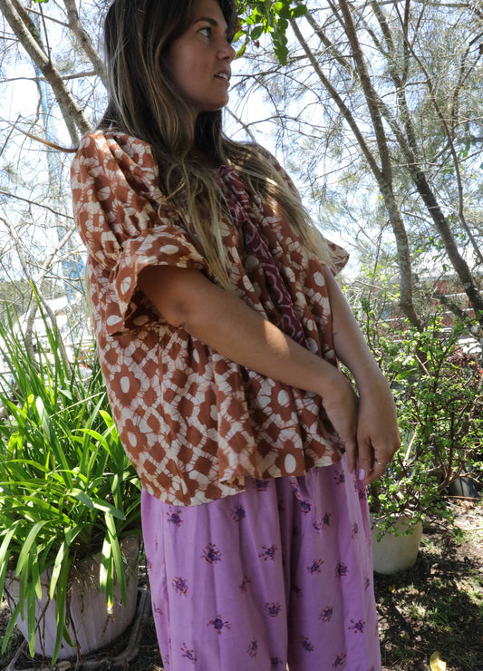 Woman wearing an ochre handblock patterned top and purple pants standing outdoors with trees and plants in the background. A perfect Christmas Fashion Outfit and Black Friday Sale
