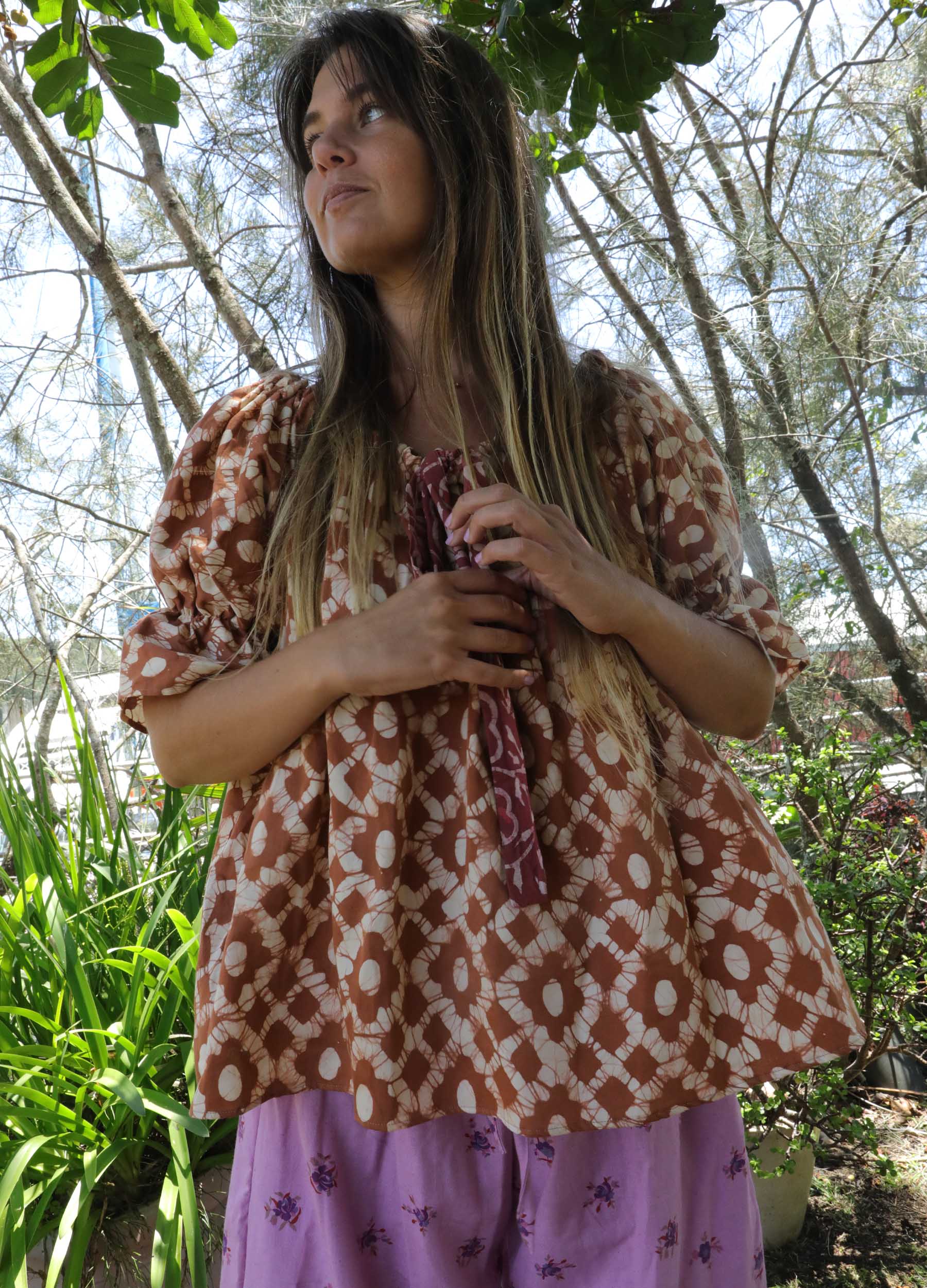 Woman wearing an ochre handblock patterned top and purple skirt standing outdoors with trees and plants in the background. A perfect Christmas Fashion Outfit and Black Friday Sale