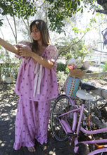 Load image into Gallery viewer, Woman in a pink outfit standing next to a pink bicycle with a basket of flowers in an outdoor setting.