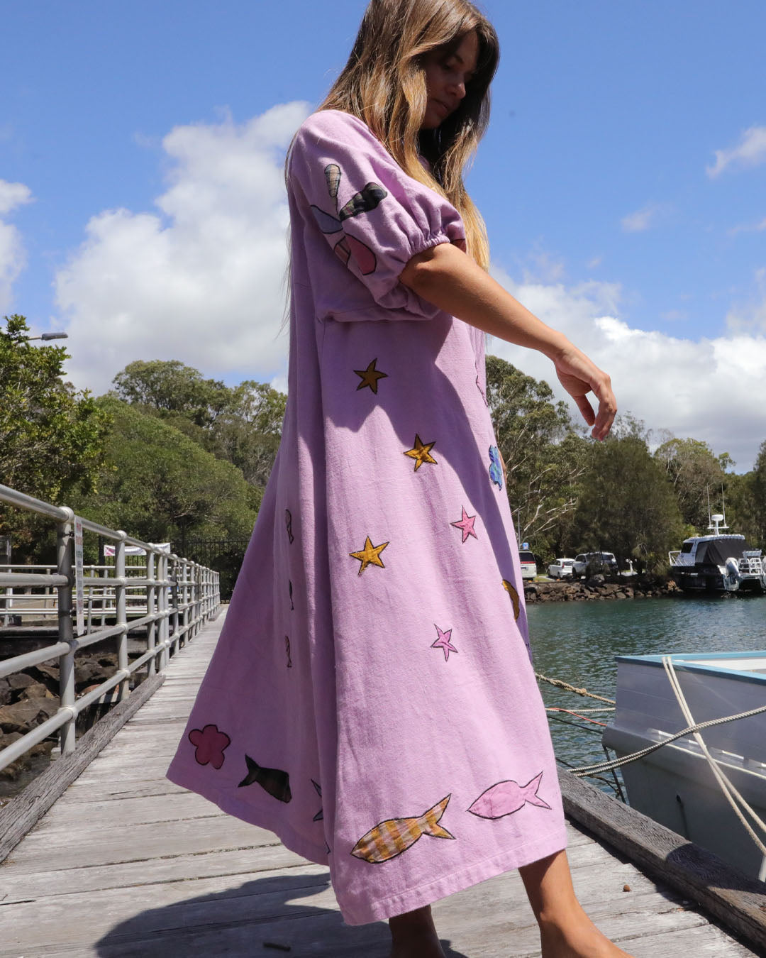 Person wearing an orchid pink dress with colorful applique embroidery standing on a dock by a body of water. One size, plus size, bohemia, Christmas fashion, Christmas Dress