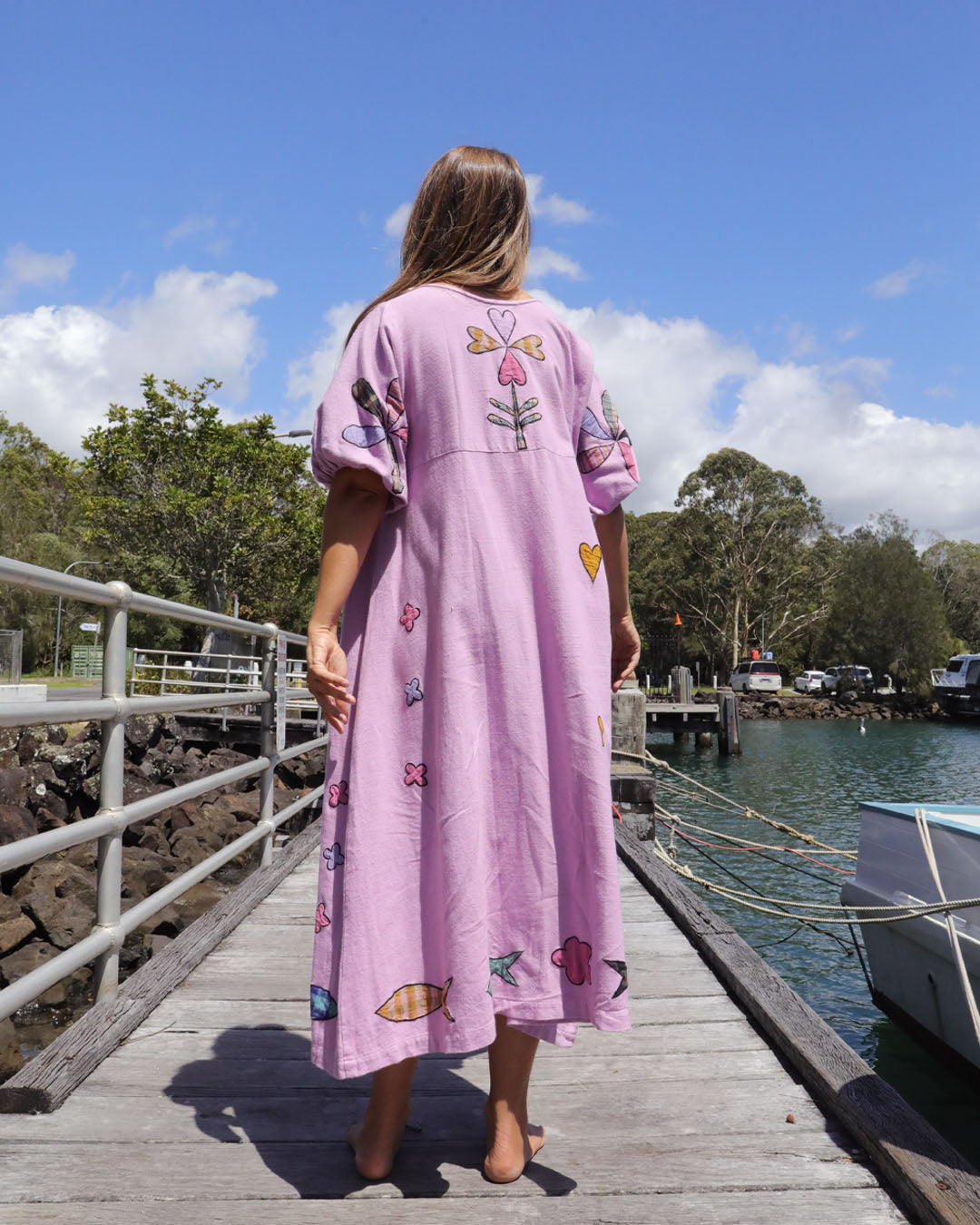 Person wearing an orchid pink dress with colorful applique embroidery standing on a dock by a body of water. One size, plus size, bohemia, Christmas fashion, Christmas Dress