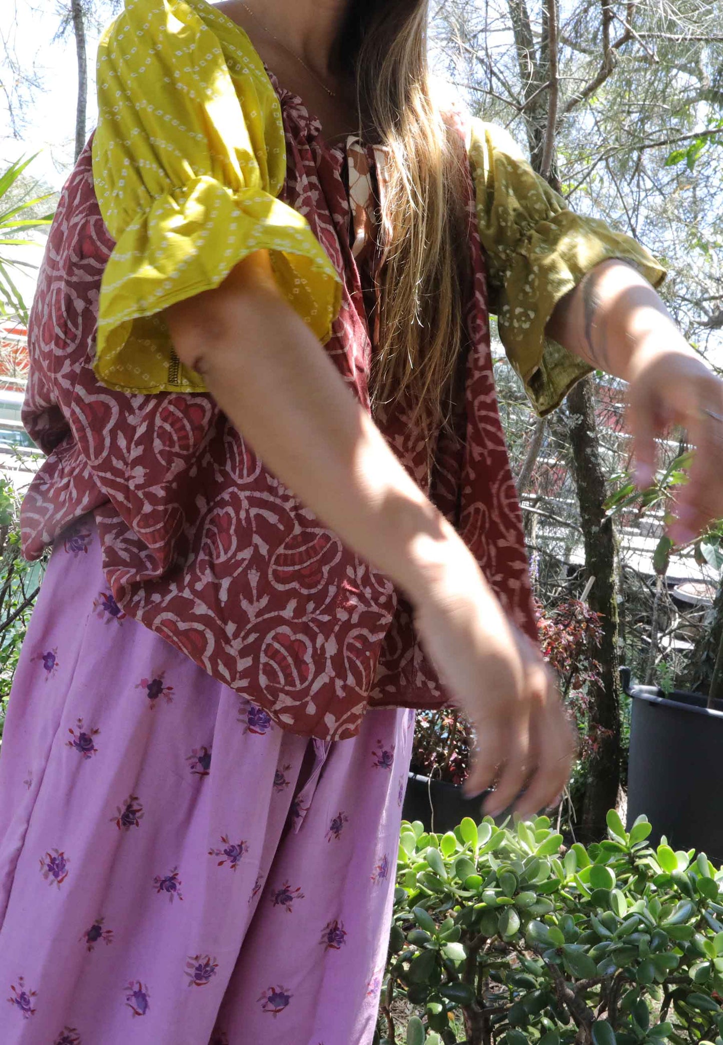 Woman wearing a pink and chartreuse handblock patterned top and purple pants standing outdoors with trees and plants in the background. A perfect Christmas Fashion Outfit and Black Friday Sale