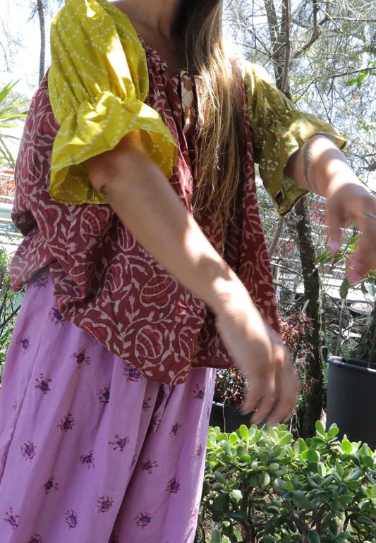 Woman wearing a pink and chartreuse handblock patterned top and purple pants standing outdoors with trees and plants in the background. A perfect Christmas Fashion Outfit and Black Friday Sale