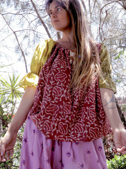 Woman wearing a pink and chartreuse handblock patterned top and purple pants standing outdoors with trees and plants in the background. A perfect Christmas Fashion Outfit and Black Friday Sale