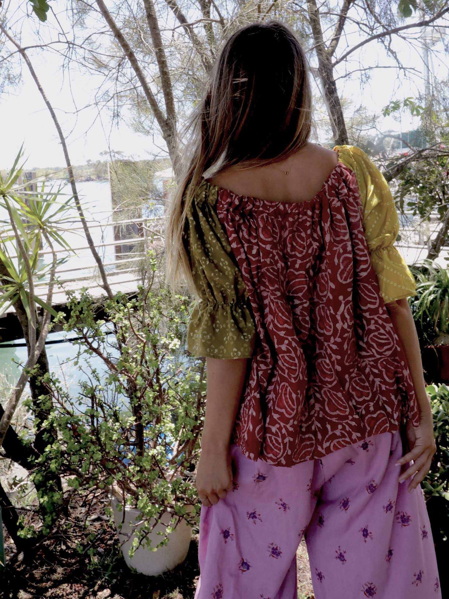 Woman wearing a pink and chartreuse handblock patterned top and purple pants standing outdoors with trees and plants in the background. A perfect Christmas Fashion Outfit and Black Friday Sale