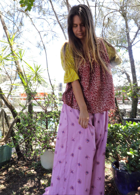 Woman wearing a pink and chartreuse handblock patterned top and purple pants standing outdoors with trees and plants in the background. A perfect Christmas Fashion Outfit and Black Friday Sale