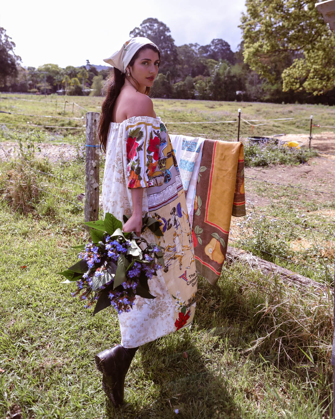 A woman stands in a grassy field wearing a cream off-the-shoulder dress made from colorful vintage-style printed fabric panels, holding a bouquet of purple flowers. A matching patterned scarf is draped over a fence behind her.