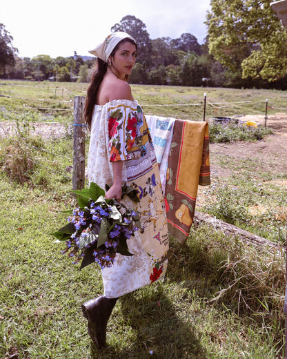 A woman stands in a grassy field wearing a cream off-the-shoulder dress made from colorful vintage-style printed fabric panels, holding a bouquet of purple flowers. A matching patterned scarf is draped over a fence behind her.