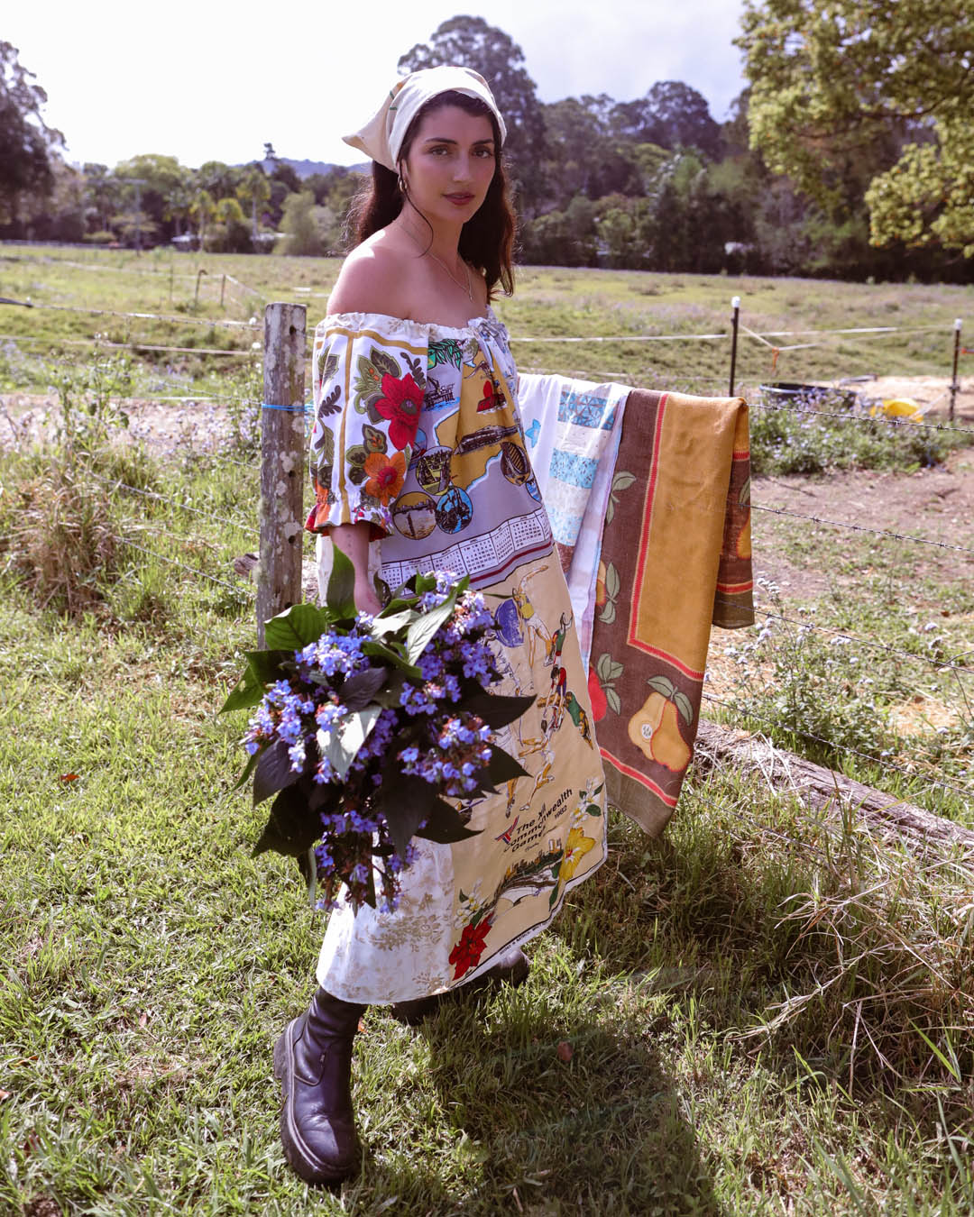 A woman stands outdoors in a grassy field wearing a bohemian-style off-the-shoulder dress with a vintage print, holding a bouquet of purple flowers. A patterned blanket is draped over a rustic wooden fence behind her.