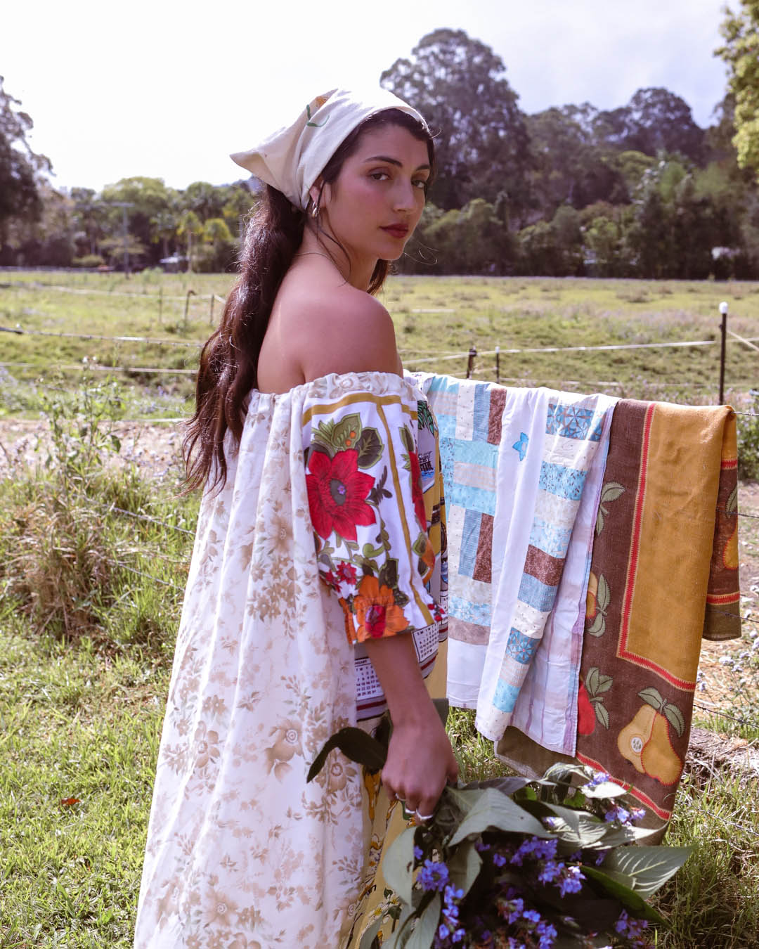 A woman outdoors in a grassy field wearing a floral off-the-shoulder dress and a headscarf, with a quilted textile draped over her shoulder. The textile features patchwork panels in blue, white, brown, and yellow with fruit and floral motifs.