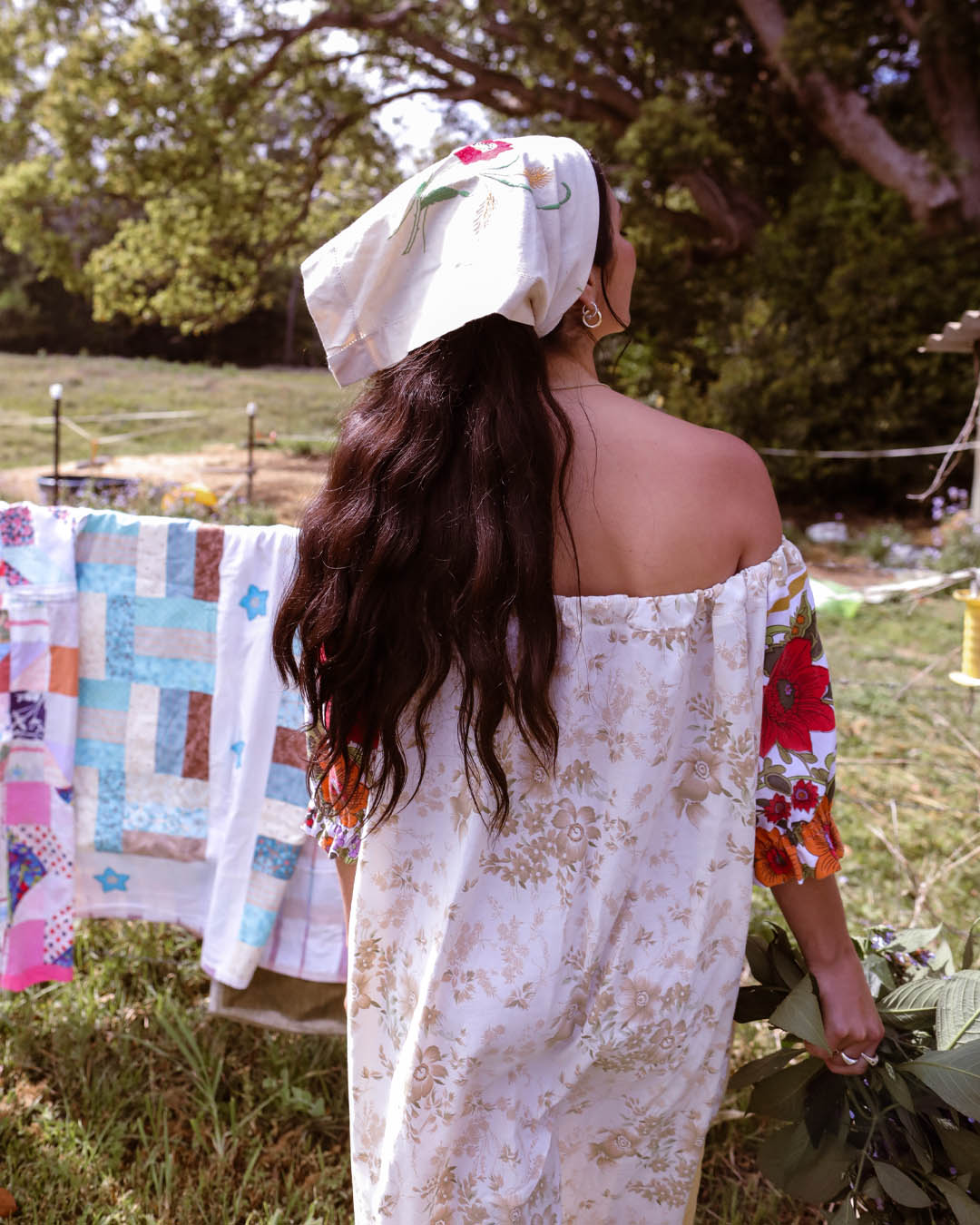 A woman viewed from behind outdoors, wearing a loose off-the-shoulder dress with a beige floral print and a white headscarf with a floral design. A quilted blanket hangs on a line in the background in a grassy outdoor setting.