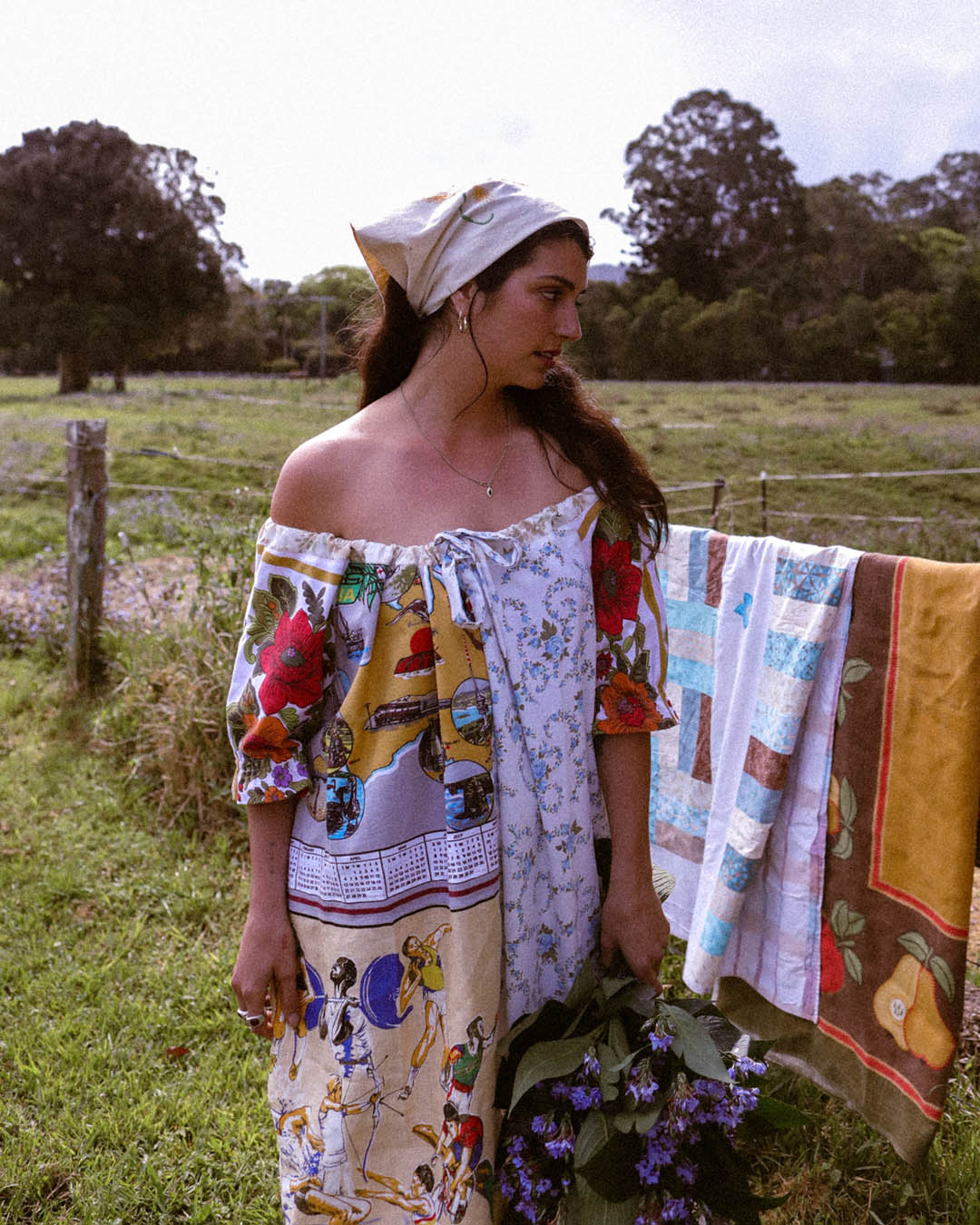 A woman stands outdoors in a grassy field wearing a patchwork off-the-shoulder dress made from colorful kitchen towels and linens, with a matching patterned headscarf. A quilted textile hangs on a fence behind her, and flowers are held in front of her.