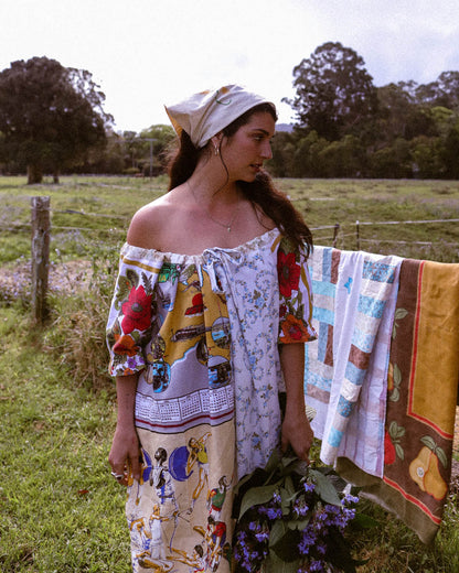 A woman stands outdoors in a grassy field wearing a patchwork off-the-shoulder dress made from colorful kitchen towels and linens, with a matching patterned headscarf. A quilted textile hangs on a fence behind her, and flowers are held in front of her.