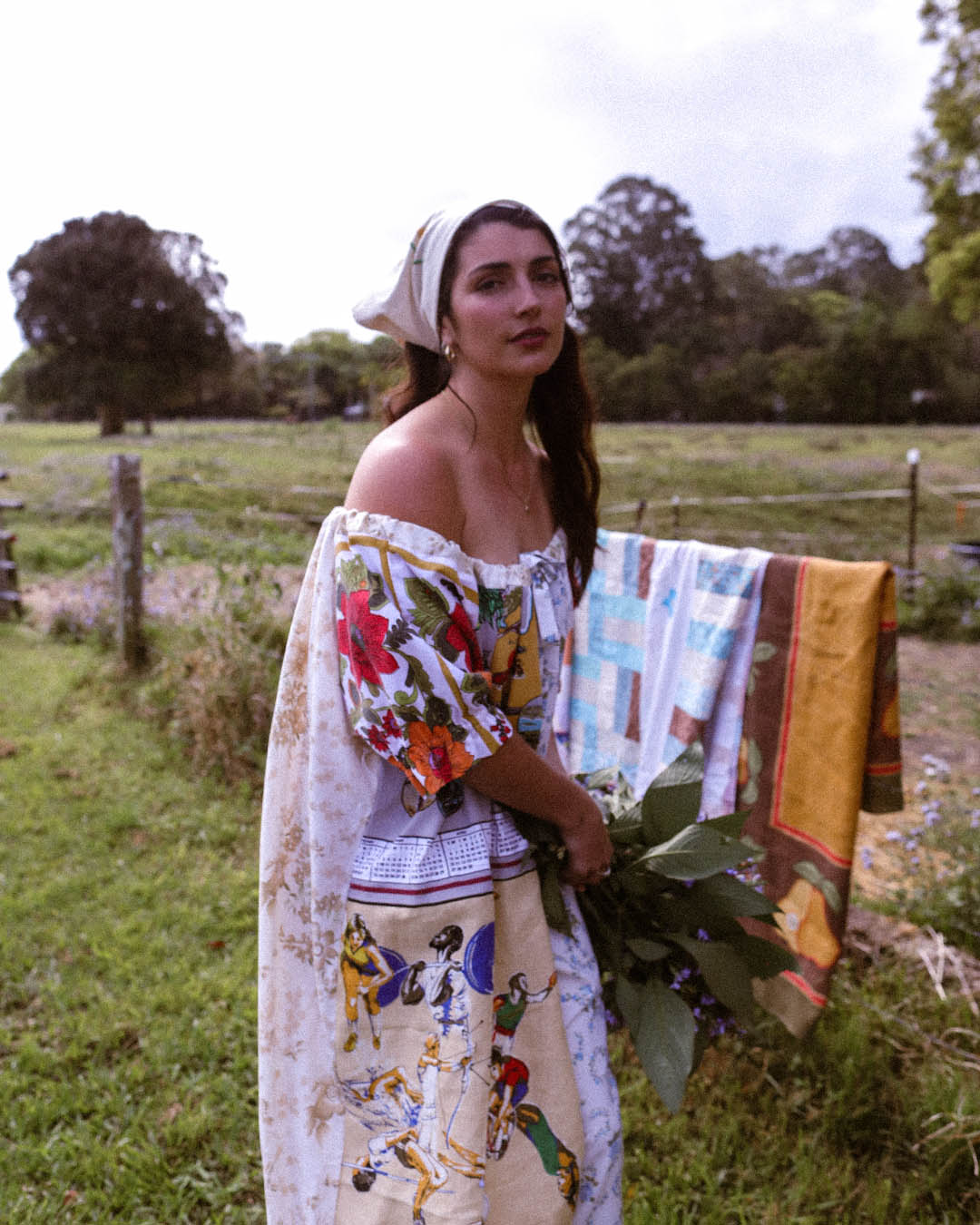 A woman stands outdoors in a grassy field wearing a colorful off-the-shoulder dress with a floral and folk-art print, holding a bundle of green leaves. A patterned textile is draped over her shoulder, and trees and a fence are visible in the background.