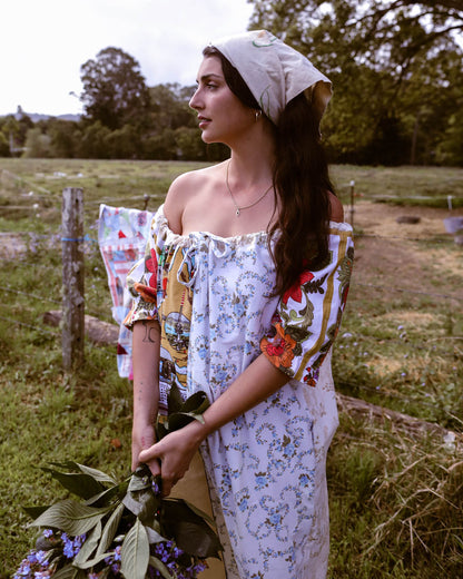 A woman stands outdoors in a grassy field wearing a white off-the-shoulder dress with colorful floral and paisley print details, a matching white headscarf, and holding green leaves and purple flowers.