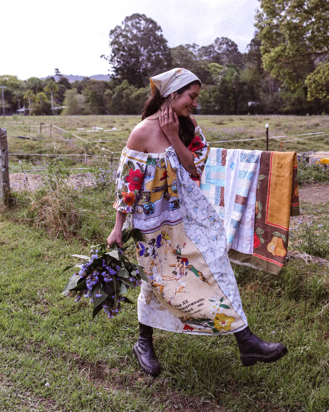 A woman walking outdoors in a grassy field wearing a colorful off-the-shoulder patchwork dress with a matching headscarf, holding a bouquet of purple flowers, with a quilt hanging on a fence behind her.