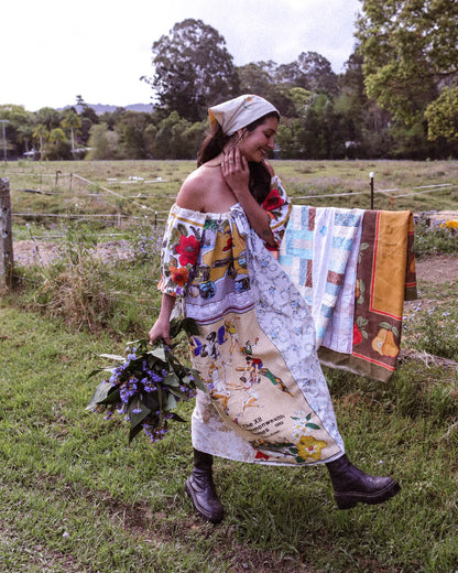 A woman walking outdoors in a grassy field wearing a colorful off-the-shoulder patchwork dress with a matching headscarf, holding a bouquet of purple flowers, with a quilt hanging on a fence behind her.